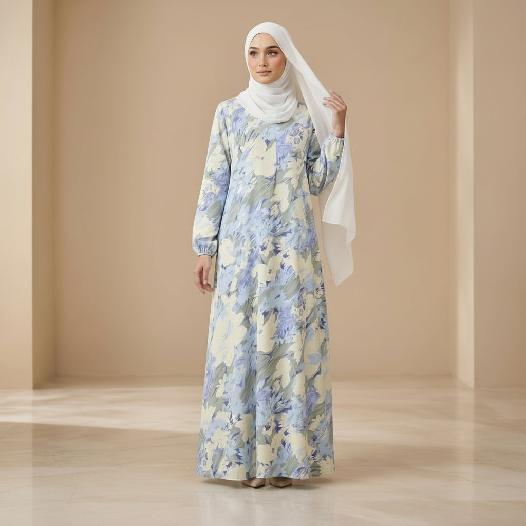 A woman modeling a long-sleeved maxi abaya with a light blue and cream floral pattern, paired with a white hijab, standing in a beige studio setting.