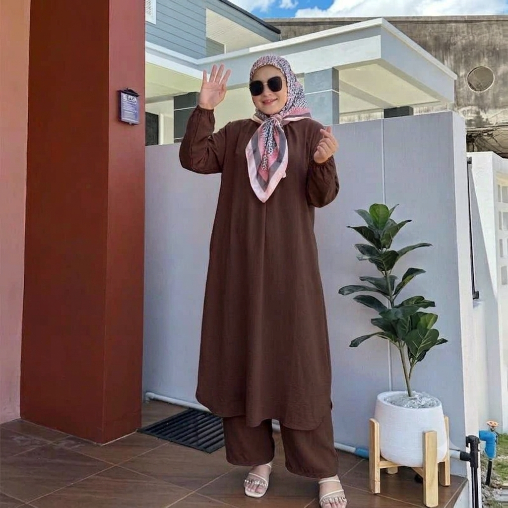 A woman wearing a brown long-sleeve modest dress with matching wide-leg pants and a pink patterned hijab stands outdoors in front of a modern house wall, waving to the camera.